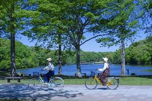 木々に囲まれた池の前を2人の女性が自転車に乗りサイクリングしている写真