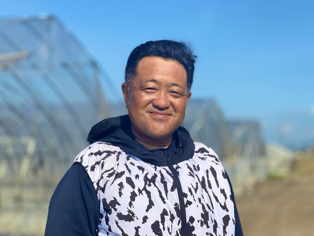 塚田さんが農地の前でカメラ目線でほほ笑んでいる