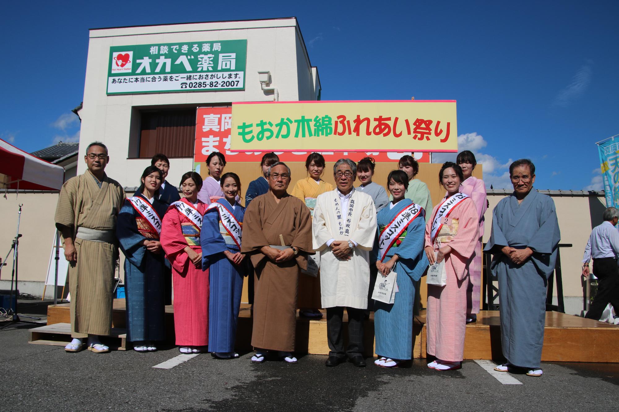 「もおか木綿ふれあい祭り」の看板の下で着物を着た人たちが並んで記念撮影を行っている写真