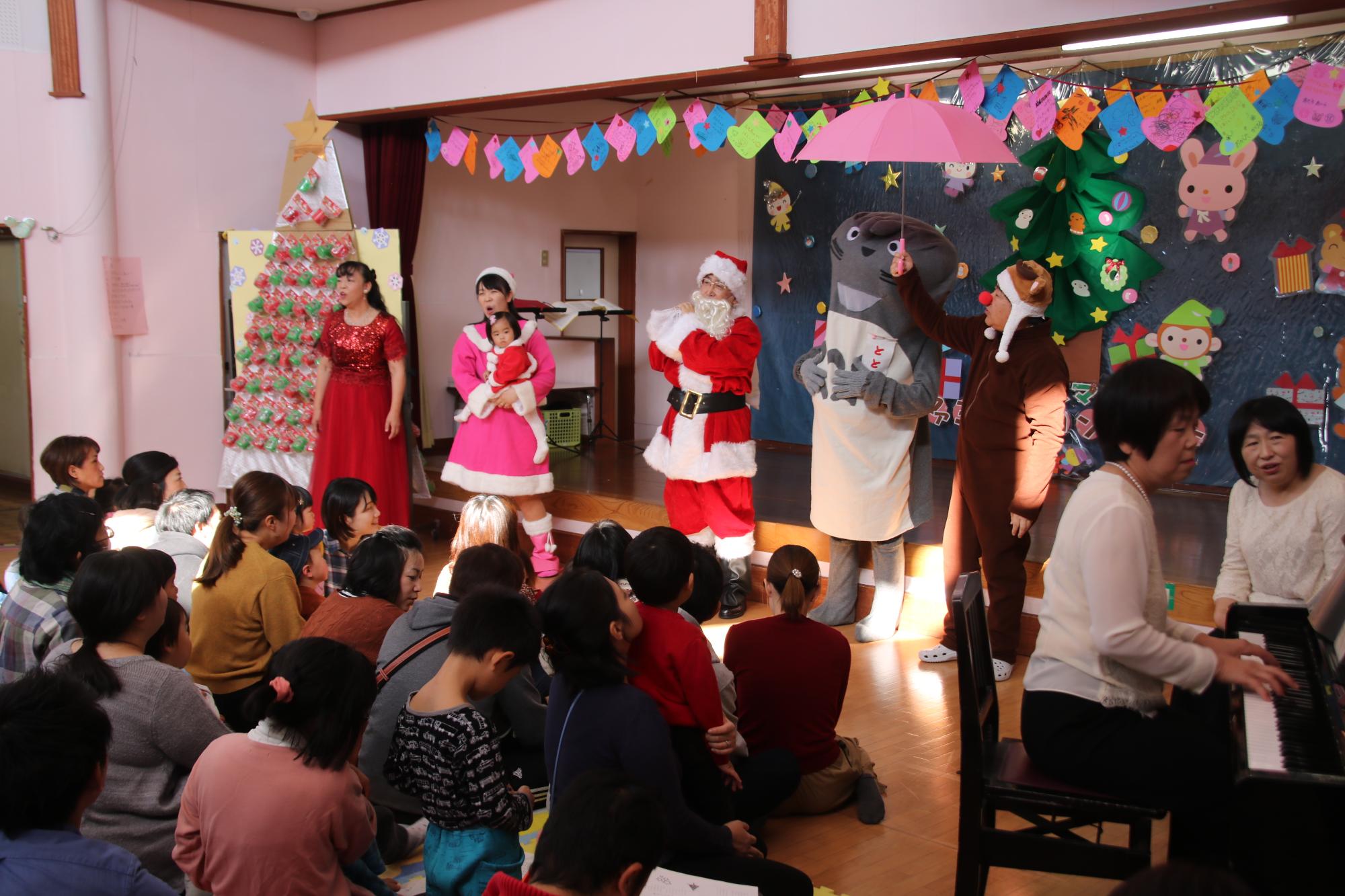 サンタクロースやトナカイなどの衣装を着た人たちが親子の前で歌を披露している写真