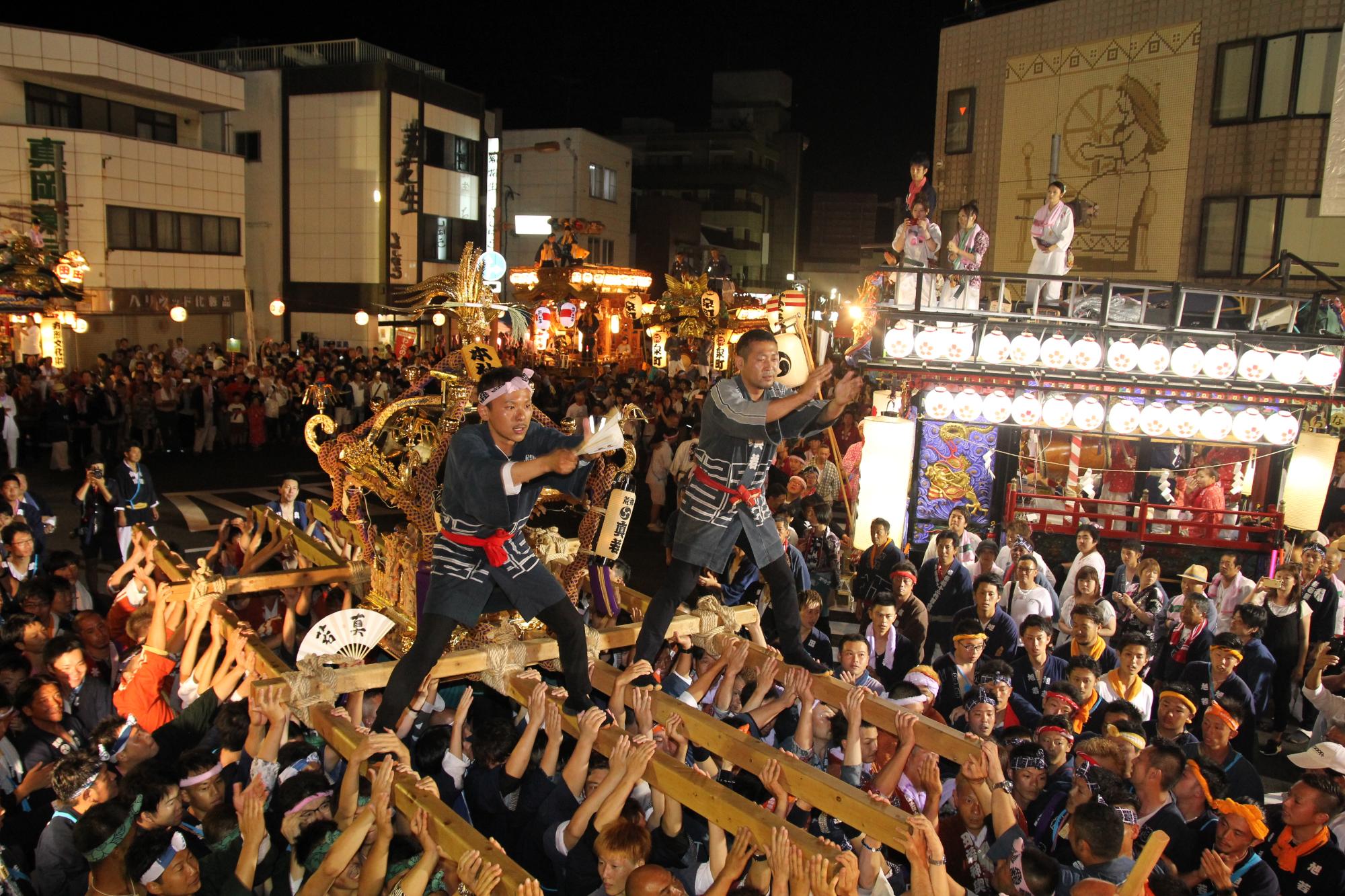 お神輿の上に2名の男性が乗りにぎわっているお祭りの写真