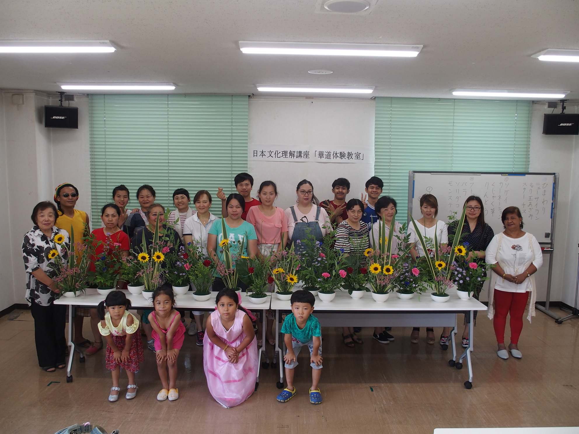 完成した生け花が並べられた台の後ろに並んで参加者達が記念撮影をしている写真