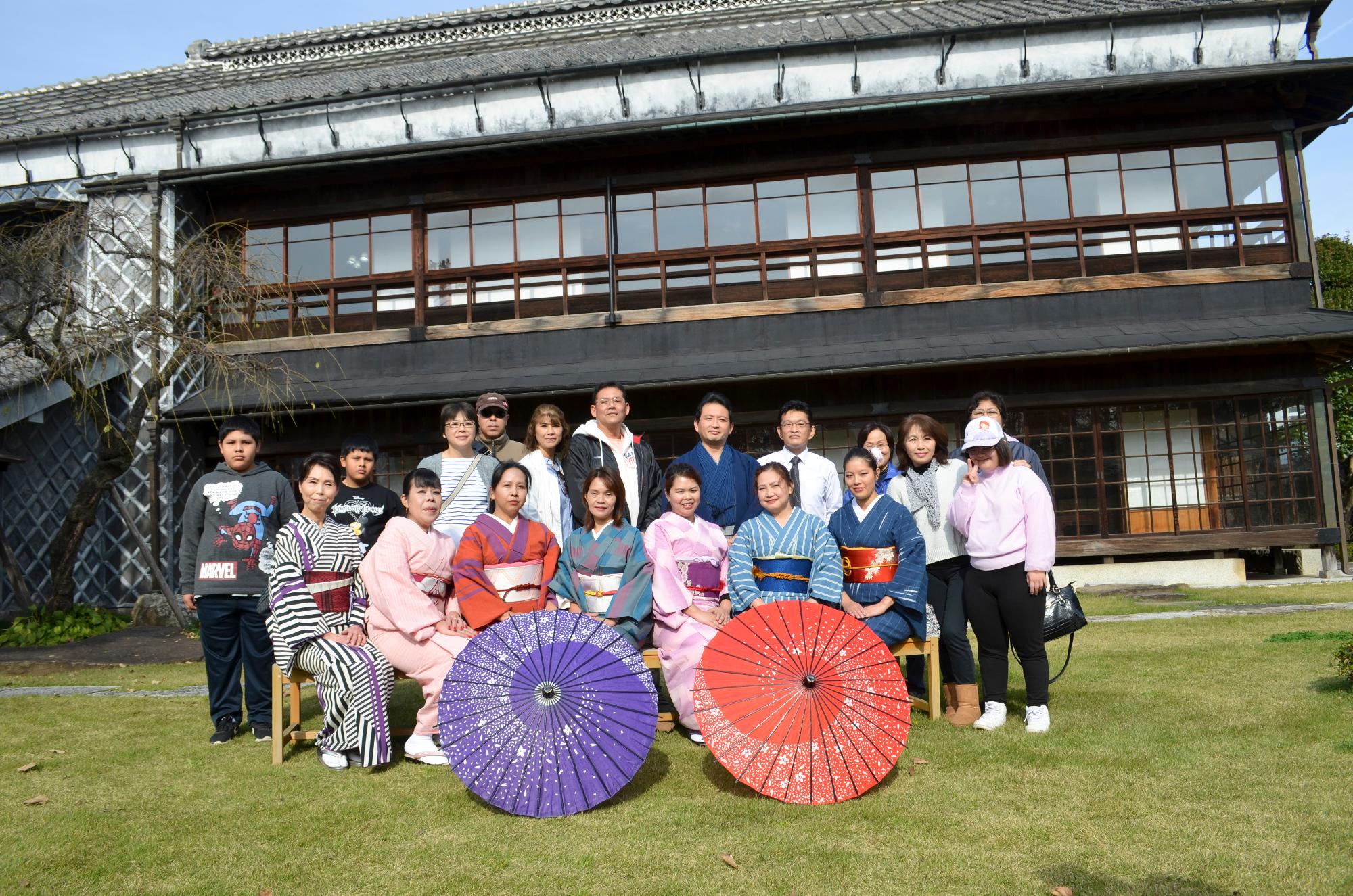 大きな2階建ての日本家屋を背景に、着物を着た参加者達が2つの和傘が置かれた後ろで集合写真を撮っている写真