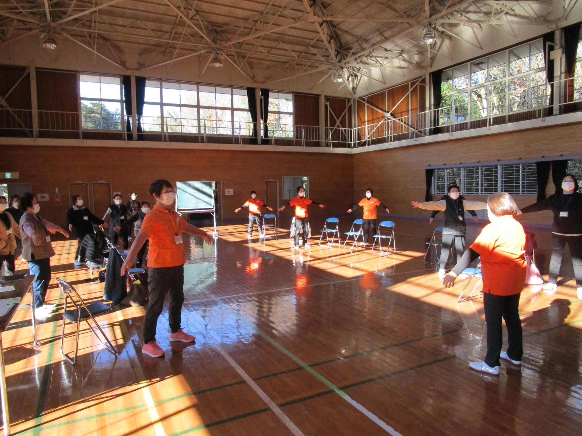 体育館の中で、参加者の方々がが間隔をあけて広がり、両手を横に広げて体操をしている様子の写真