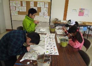 テーブルの上で4人の大人と子どもたちが、野鳥の木型に絵の具で色を塗っている写真です。