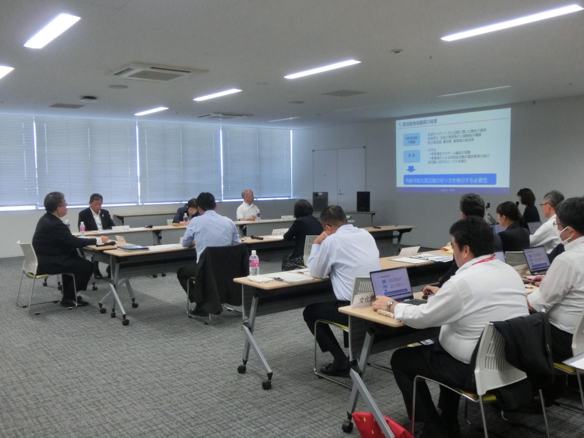 プロジェクターに映像を写しながら、マイクを持った女性が話をしており、参加者の方々が席について、資料に目を通しながら話を聞いている、令和6年度第1回真岡市総合教育会議の様子の写真