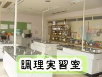 調理台がならんでおり、右側の壁側に調理器具が置かれた棚が設置されている調理実習室の写真