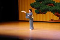 舞台の松の木の背景をバックに、着物を着た女性が扇子を持ち踊りを披露している写真
