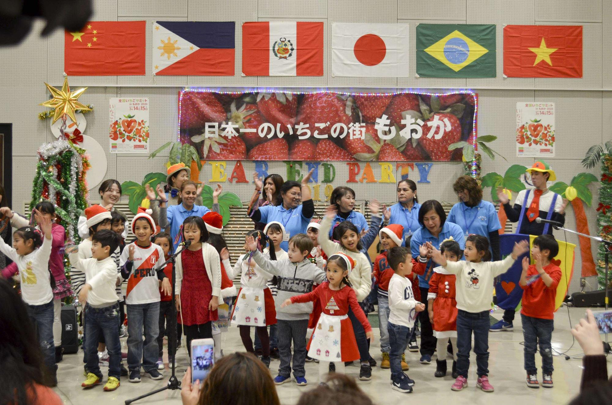 中国、フィリピン、ペルー、日本、ブラジル、ベトナムの国旗が掲げられた会場で、赤と白の服を着て、サンタの帽子を被った子供たちが発表をしている様子の写真