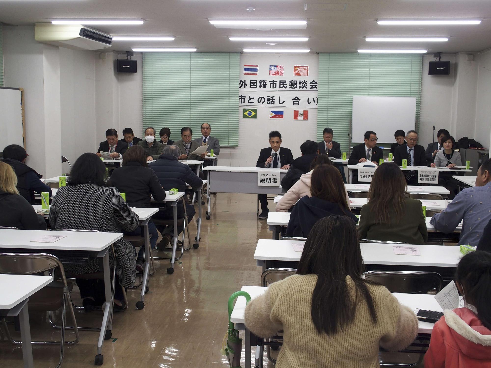 6か国の国旗と「外国籍市民懇談会 市との話し合い」と書かれた紙が壁に貼られた室内で参加者達が席について懇談会が行われている様子の写真