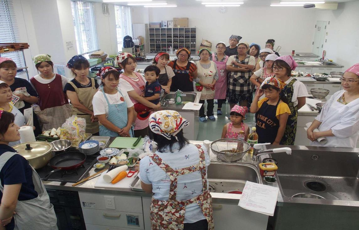 1つの調理台の周りに参加者達が集まり、調理台で作業をしている女性を見ている写真