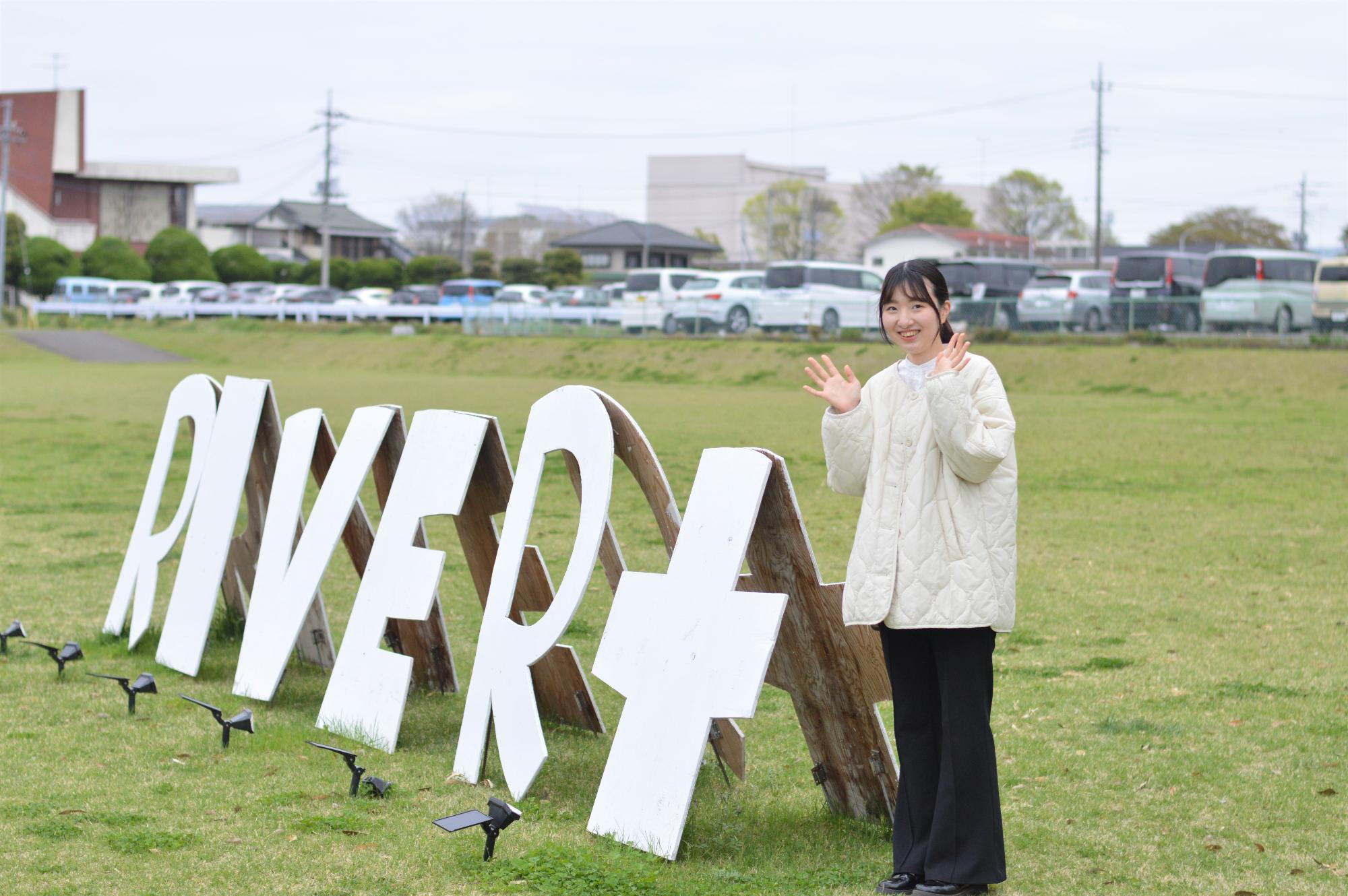 RIVER＋の看板の横に立っている笑顔の岩崎さんの写真