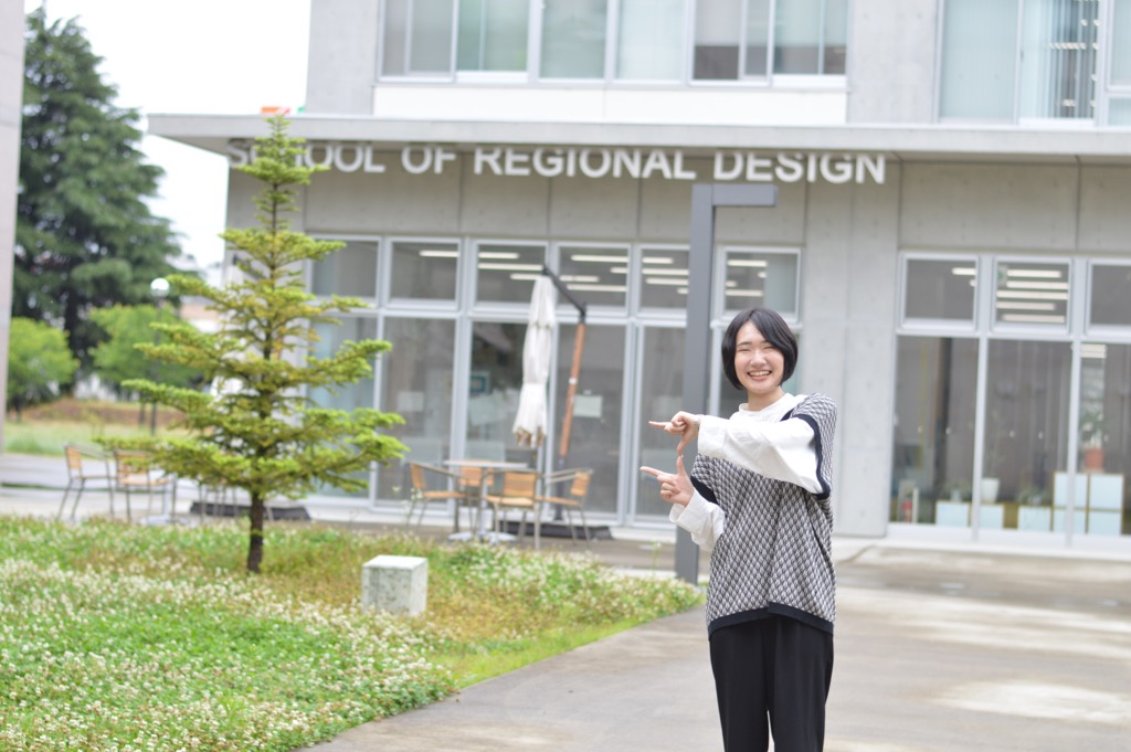 SCHOOL OF REGIONAL DESIGNと書かれた建物の前でポーズをとって笑顔で写る石川 すずさんの写真