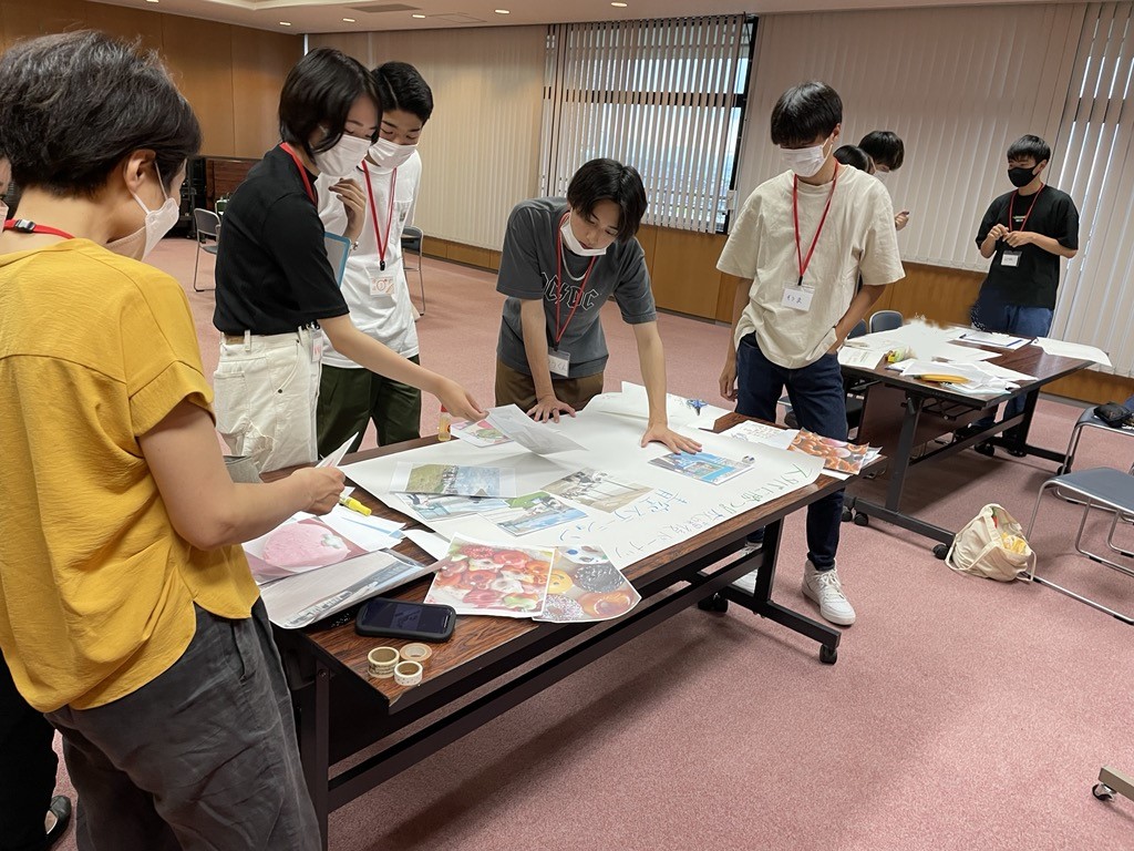 長机の周りに5名の参加者が集まり、模造紙に企画をまとめている様子の写真