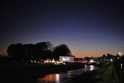 オレンジ色が少し残る夜空の下、イベント会場の灯りや映画上映されているスクリーンの光が会場横の川の水面に反射している写真