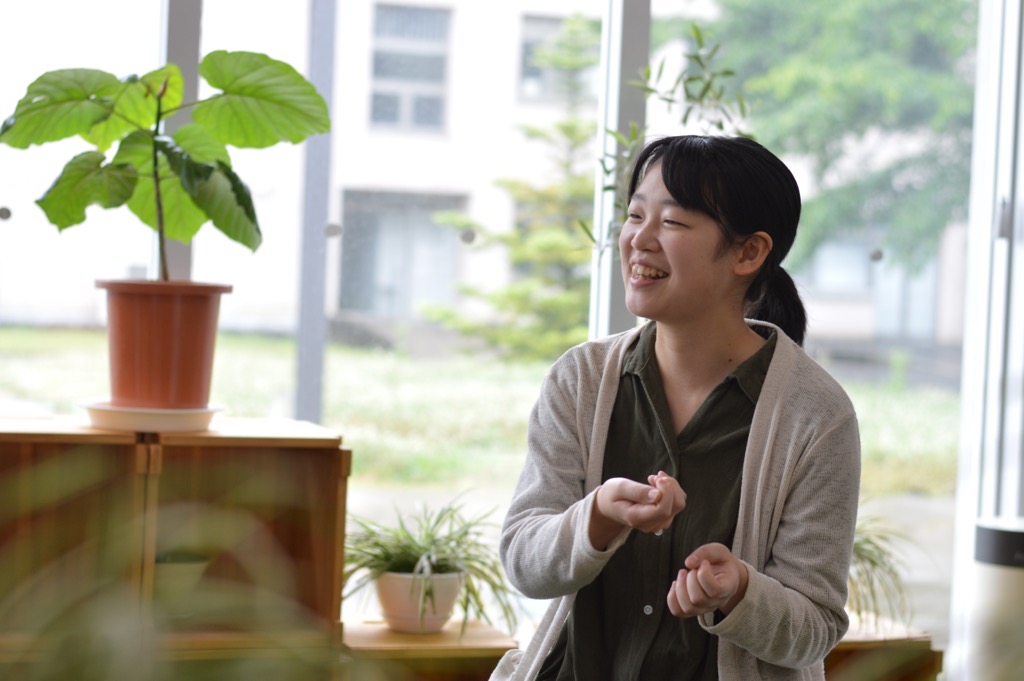 鉢植えの植物がある室内で笑顔で座っている阿藤 結衣さんの写真