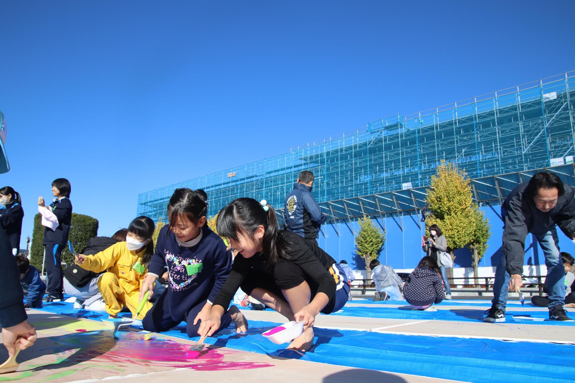 建設中の複合交流拠点の前で大きなベニヤ板のキャンバスに絵を描く子供たち