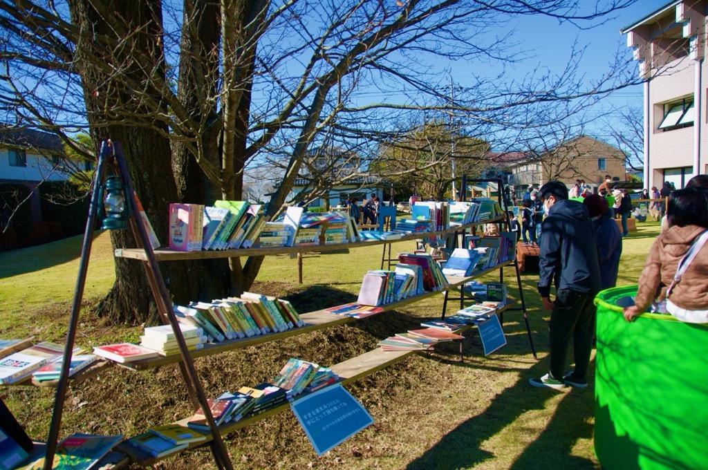 大きな木の前に置かれた本棚に沢山の本が並べられている青空図書館の写真