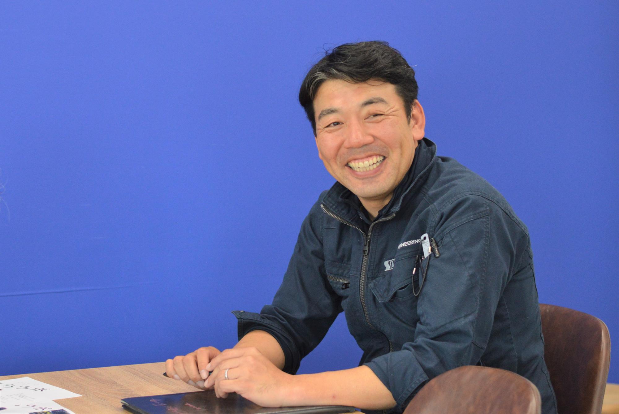 青い壁を背景に椅子に座り笑顔を見せている伊澤 学さんの写真