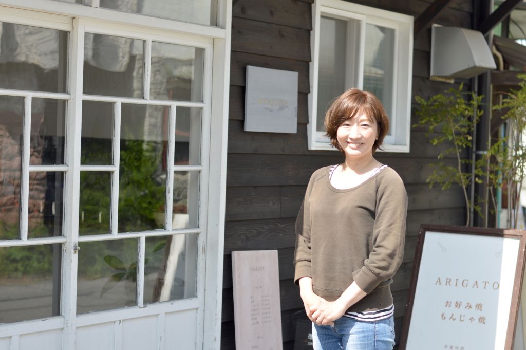 お好み焼きARIGATOの入り口前で両手を前に添えて笑顔で立っている久保田 有紀さんの写真