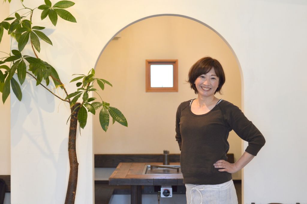 左側に植物があるアーチ形の入り口前に笑顔で立っている久保田有紀さんの写真