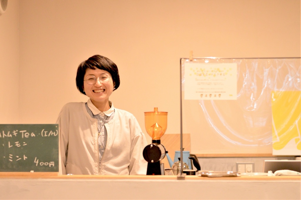 ドリンクメニューが置かれたカウンター奥にいる笑顔の岡 綾さんの写真