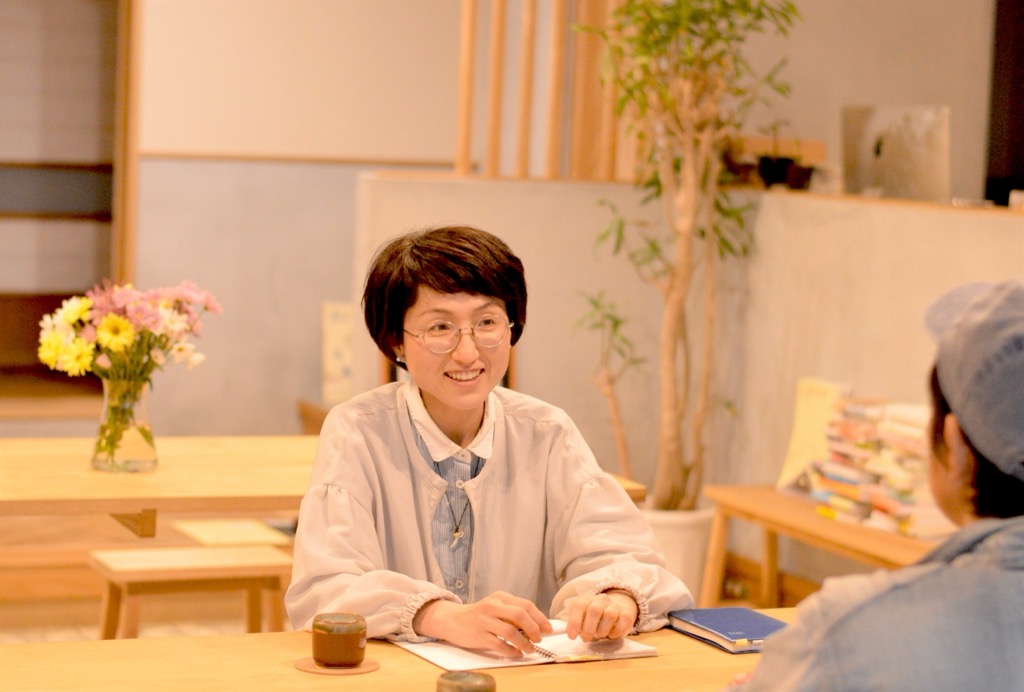 丸い眼鏡をかけ、インタビューを受けている岡 綾さんの写真