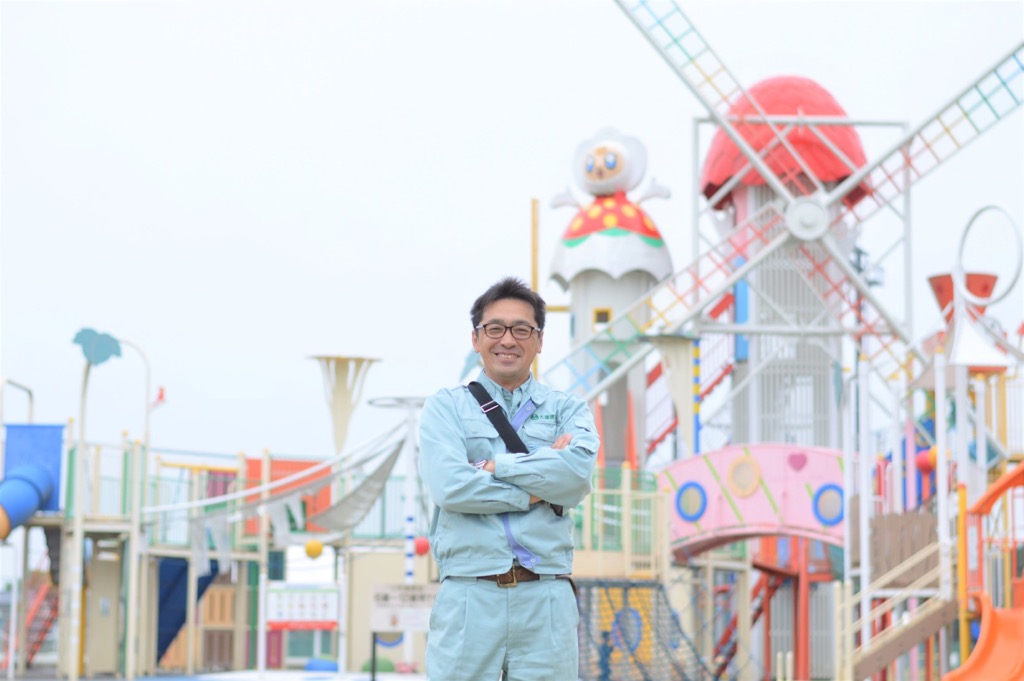 風車のような形をした大きなコンビネーション遊具の前で、両腕を組んで笑顔で立っている大瀧武志さんの写真