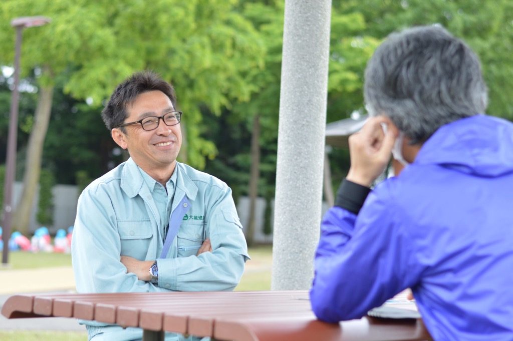 屋外のテーブルに男性の方と向き合って座り笑顔を見せている大瀧 武志さんの写真