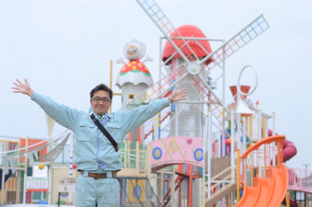 滑り台などが付いた大きなコンビネーション遊具の前で両手を上に広げて立っている大瀧 武志さんの写真
