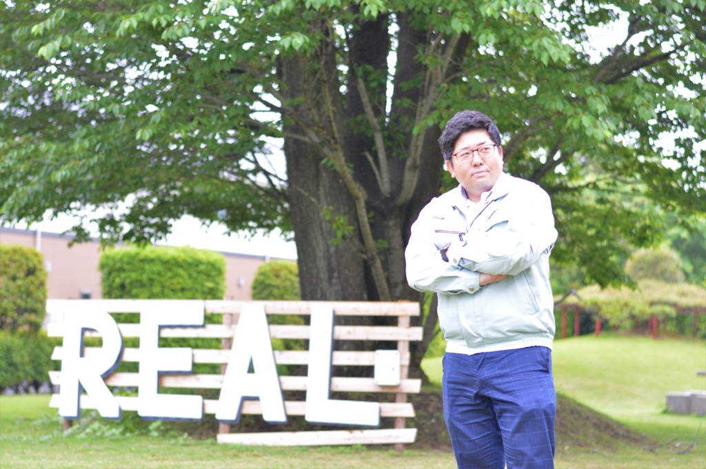 REALと書かれた木製の看板の前で両腕を組んで遠くを見ている上澤さんの写真